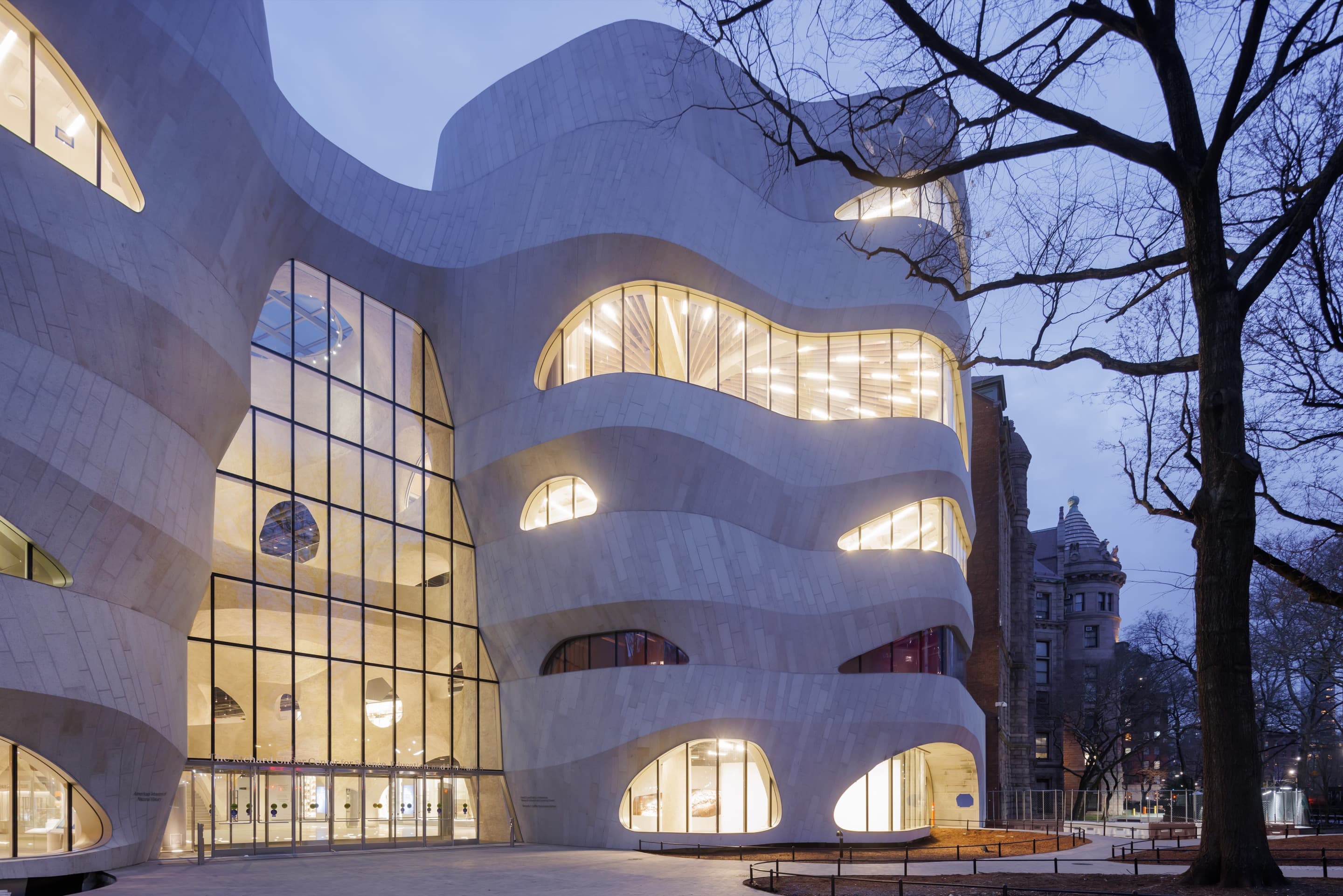 Gilder Center at the American Museum of Natural History at dusk. The building has an undulating facade and large, organically-shaped windows.