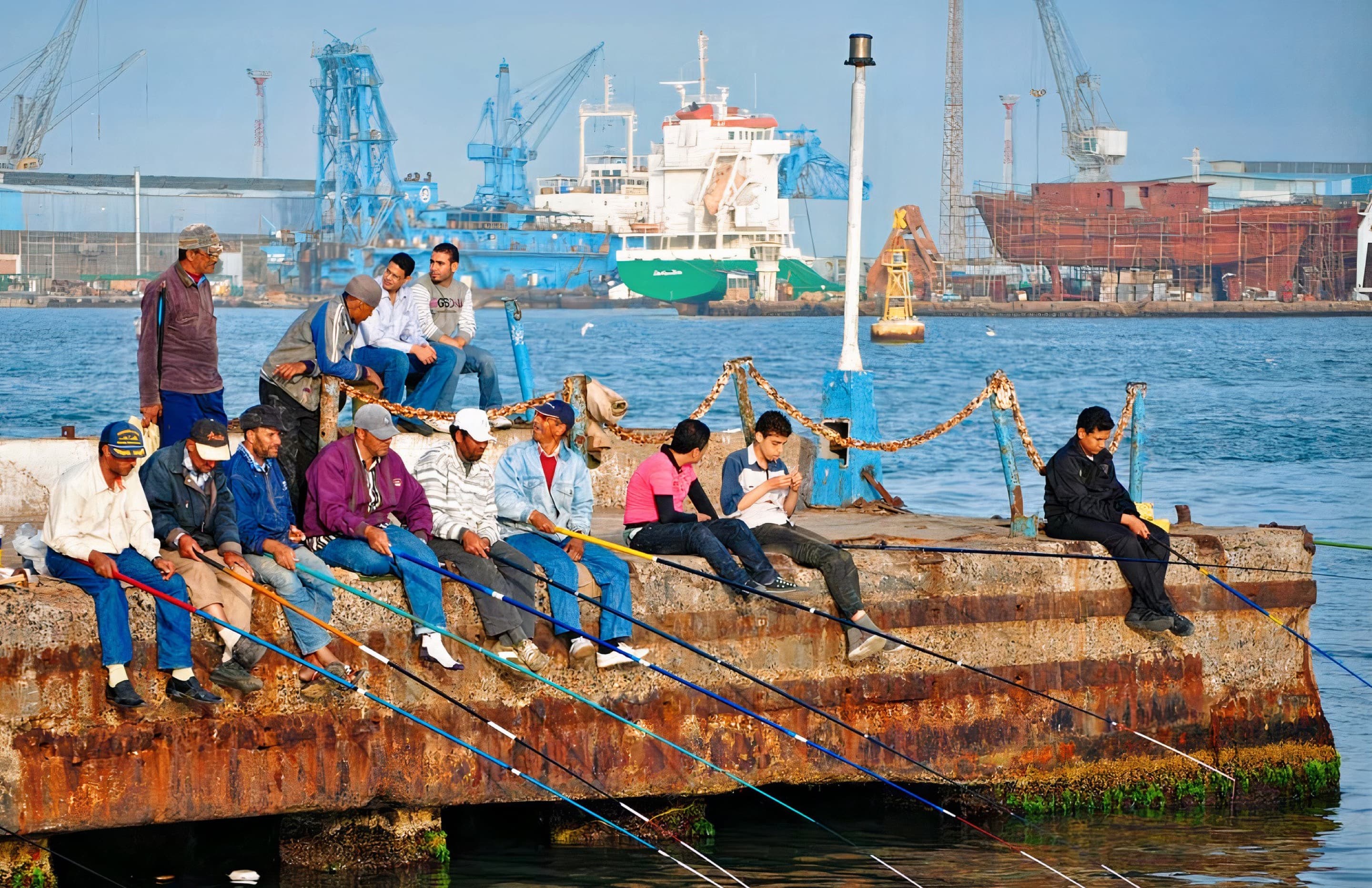 Fishermen sit with their rods down along the dock at Port Said, Egypt. There are crate cranes and freight ships in the background.